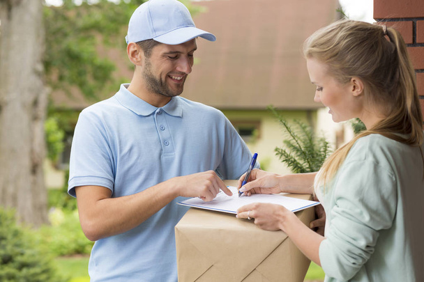 Gelukkig courier in blauwe uniform en vrouw ondertekening na ontvangst van de levering van het pakket - Foto, afbeelding