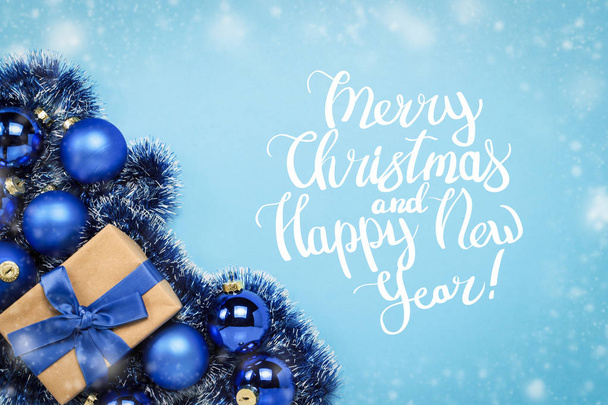 Gift box with blue ribbon, blue Christmas tree toys, balls on a blue background with falling snow. Added text Merry Christmas and Happy New Year. Flat lay, top view. - Photo, Image