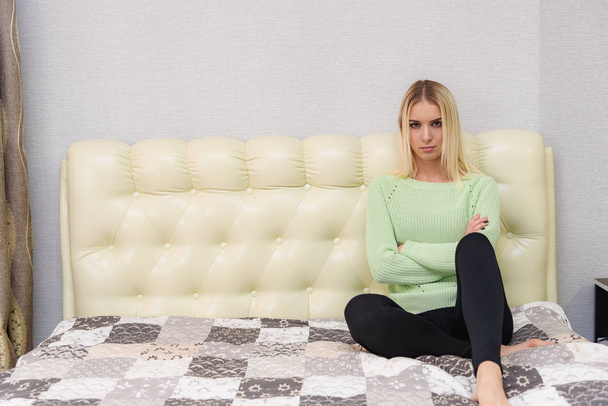 Photo portrait of a beautiful blonde girl in a room sitting on the sofa. She is right in front of the camera, looks serious. - Valokuva, kuva