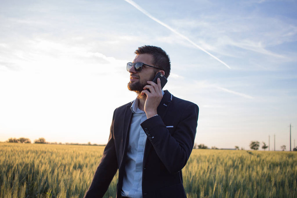 Young man in black suit speacking on phone in wheat field, during beautiful sunset - Valokuva, kuva