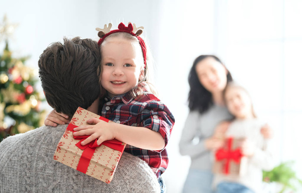 Счастливого Рождества и Рождества! Мама, папа и дети обмениваются подарками. Родители и дочери веселятся возле деревьев в помещении. Любимая семья с подарками в комнате
. - Фото, изображение