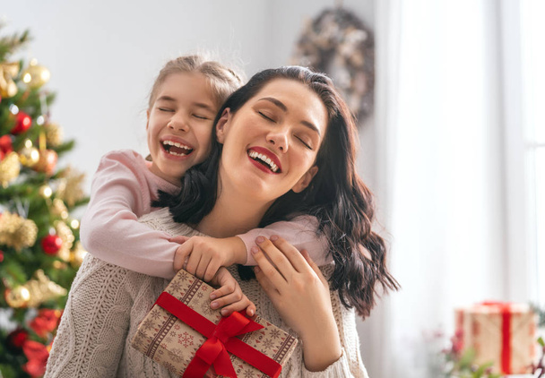 Merry Christmas and Happy Holidays! Cheerful mom and her cute daughter girl exchanging gifts. Parent and little child having fun near tree indoors. Loving family with presents in room. - Foto, Imagem