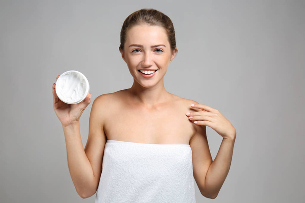 Beautiful young woman wrapped in towel holding jar of cream on grey background - Photo, Image