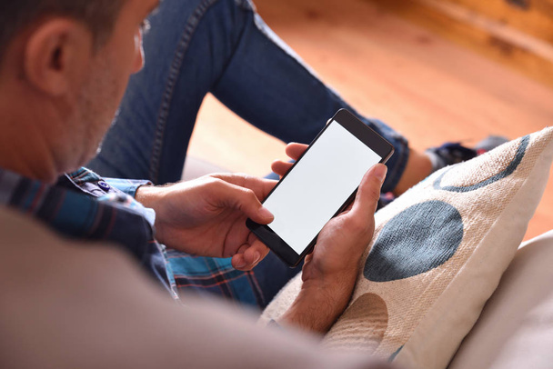 Adult man with blue shirt and jeans sitting on a sofa using a smartphone. Raised viewpoint - Foto, imagen