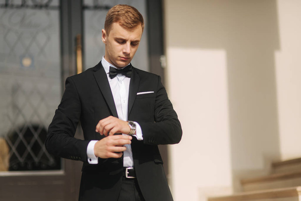 Fashion groom in trendy suit posing to photographer. Blond hair man with bow tie and other accsessories stand by the beautiful hotel - Fotó, kép