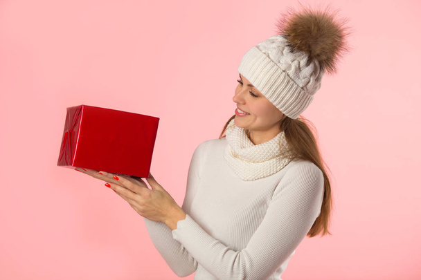 όμορφη νεαρή κοπέλα στο χειμερινό καπέλο με ένα δώρο στο χέρι σε ροζ φόντο - Φωτογραφία, εικόνα