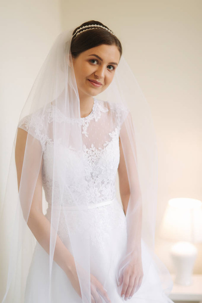 schöne Braut steht am Fenster in weißem Hochzeitskleid. Charmante Dame - Foto, Bild