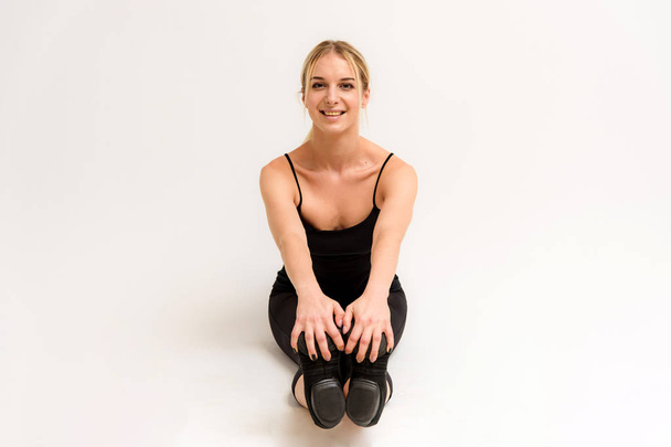 Studio photo of a beautiful blonde girl doing fitness stretching exercises on a white background. She is right in front of the camera, smiling and looking happy. - Фото, зображення