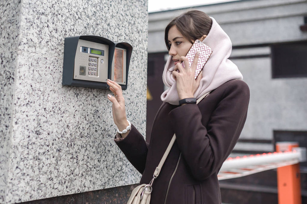 brunette woman calls into the intercom on the street, autumn day in warm clothes - Photo, image