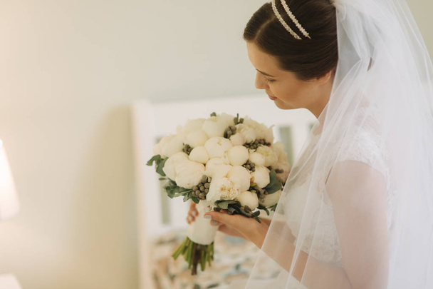 Beautiful and tenderness bride in white dress and veil. Hold bouquet of flowers in hands - Photo, Image