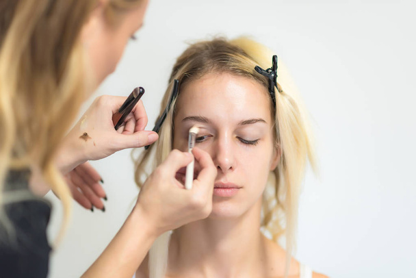 The work of a professional makeup artist - beautician, makes makeup with a brush on the face of a beautiful blonde with shadows on the eyes of the model. Painting of eyebrows. - Фото, изображение