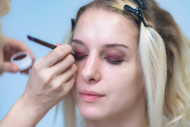 The work of a professional makeup artist - beautician, makes makeup with a brush on the face of a beautiful blonde with shadows on the eyes of the model. Painting of eyebrows. - Photo, image