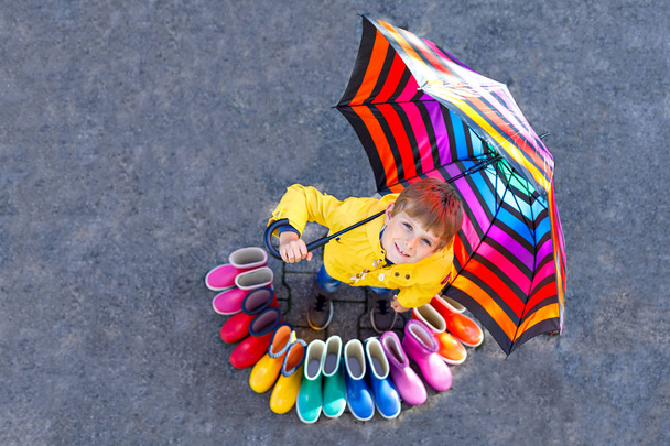 Little kid boy and group of colorful rain boots. Blond child standing under umbrella. Close-up of schoolkid and different rubber boots from high angle. Footwear for rainy fall - Foto, Imagen