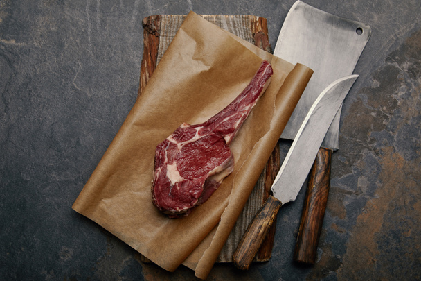 top view of raw rib eye steak on baking paper with kitchen knives on grey background - Fotoğraf, Görsel