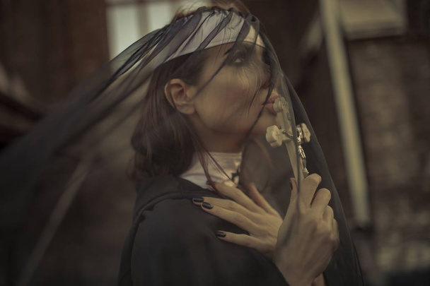 Portrait de la jeune religieuse en prière en soutane noire, manteau et cape transparente avec croix dans les mains. Vue latérale
. - Photo, image