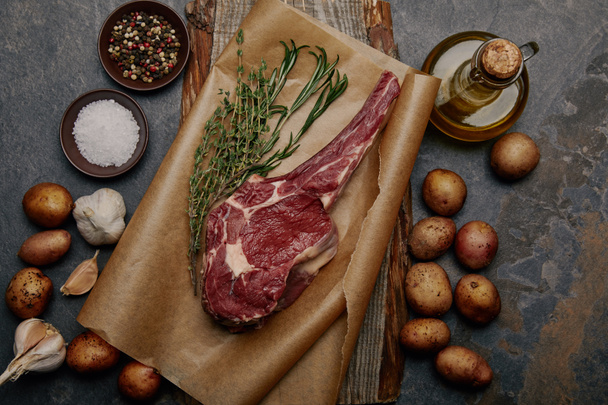 ruwe rib eye steak op bakken papier met specerijen, kruiden en aardappelen op grijze achtergrond - Foto, afbeelding