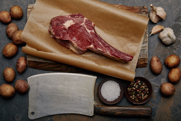 flat lay with raw rib eye steak on baking paper with butcher knife, spices, potatoes and garlic - Фото, зображення