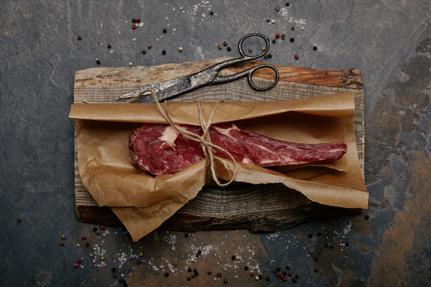 bovenaanzicht van ruwe rib eye steak verpakt in bakken papier op een houten bord met kruiden op grijze achtergrond - Foto, afbeelding