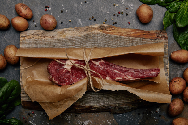 ruwe rib eye steak verpakt in bakken papier op een houten bord met specerijen en aardappelen - Foto, afbeelding