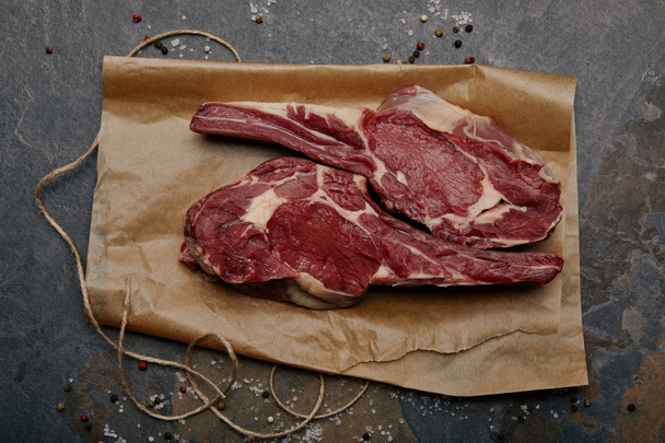 bovenaanzicht van ruwe rib eye steak op bakken papier met specerijen op grijze achtergrond - Foto, afbeelding