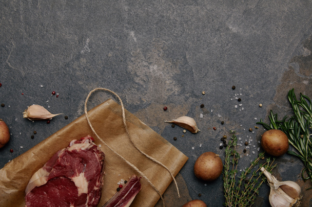 top view of raw meat steak on baking paper with spices, herbs and potatoes on grey background - Foto, immagini