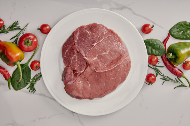 top view of fresh raw meat on plate with vegetables and herbs on white background - Photo, Image