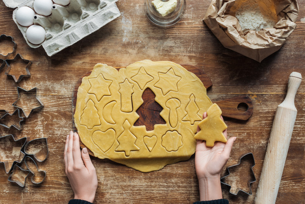 bijgesneden schot van vrouwelijke handen, ruwe deeg, ingrediënten voor christmas cookies bakkerij en cookie cutters op houten tafelblad - Foto, afbeelding