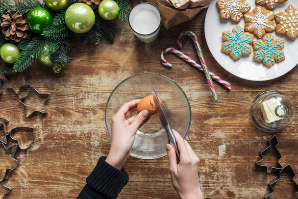 cropped shot of woman cracking chicken egg into bowl while making dough for christmas cookies on wooden tabletop with decorative wreath - Photo, Image