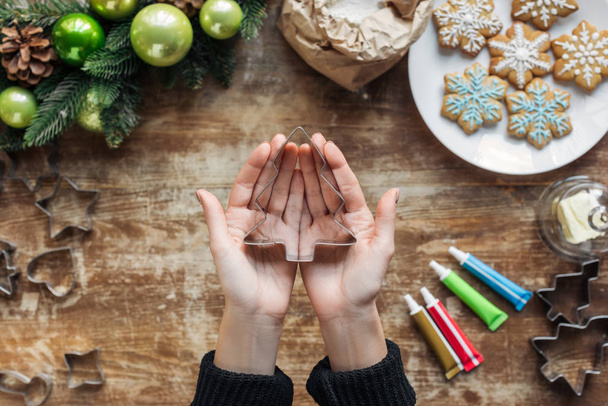 partial view of woman holding fir tree shaped cookie cutter in hands with christmas wreath and ingredients on wooden tabletop - Фото, зображення