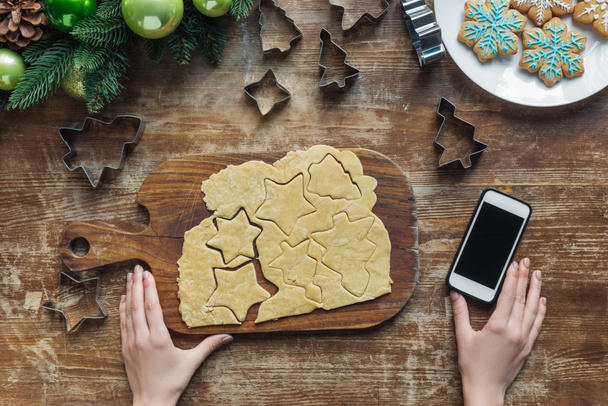 vista parcial de la mujer, teléfono inteligente con pantalla en blanco y masa cruda para galletas de Navidad en la mesa de madera
 - Foto, imagen