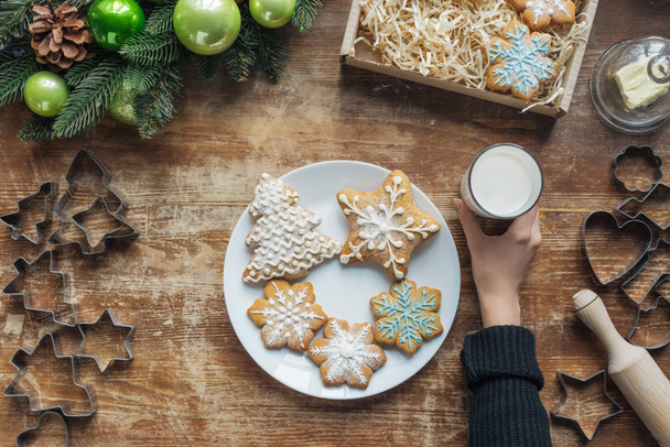 částečný pohled ženy držící sklenice mléka na dřevěnou desku s dekorativní vánoční věnec, soubory cookie a kartonovou krabici - Fotografie, Obrázek
