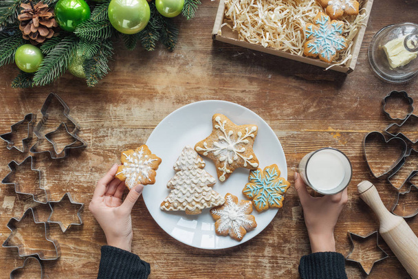 partial view of woman holding glass of milk and cookie on wooden tabletop with decorative christmas wreath and cardboard box - Foto, afbeelding