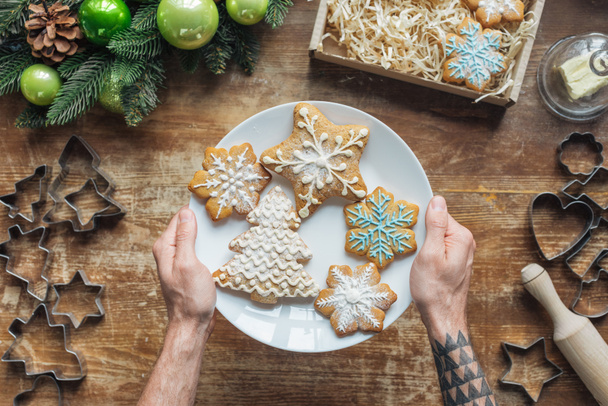 cropped shot of man holding plate with homemade christmas cookies on wooden surface - Foto, afbeelding