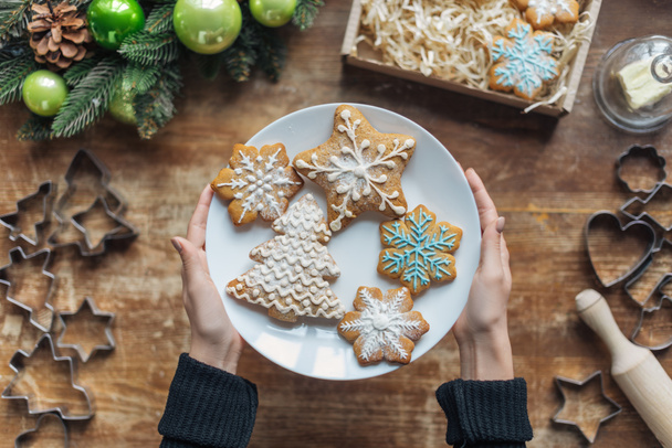 partial view of woman holding plate with homemade cookies on wooden tabletop with decorative christmas wreath and cardboard box - Foto, Bild