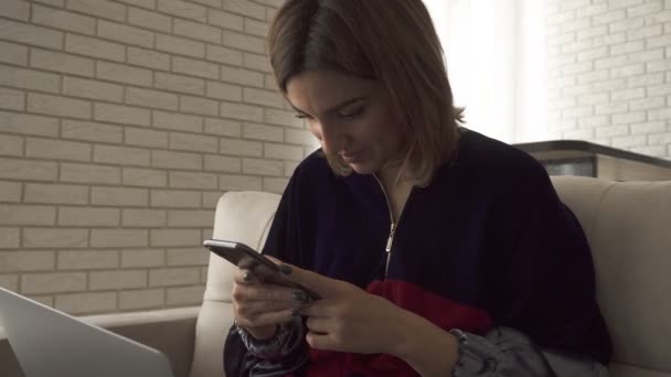A girl typing on mobile phone, then smiling and typing on a laptop. Female typing on keyboard. Freelancer girl working from home - Materiał filmowy, wideo