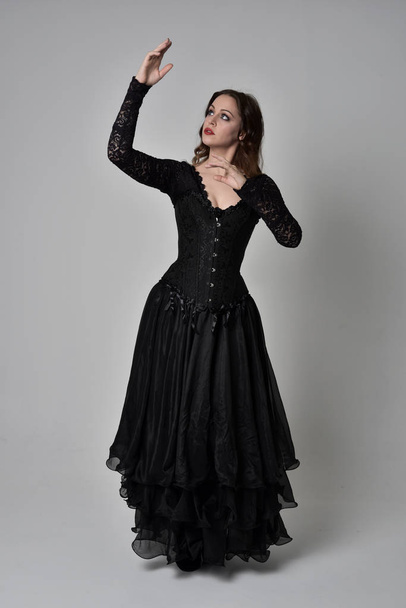 full length portrait of brunette girl wearing long black lace gown wit corset. standing pose, isolated on grey studio background. - Photo, Image