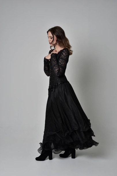 full length portrait of brunette girl wearing long black lace gown wit corset. standing pose, isolated on grey studio background. - Fotó, kép