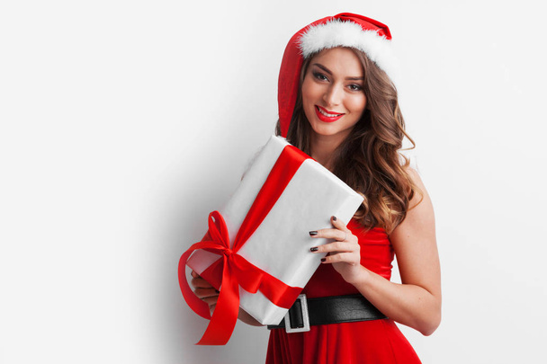 Beautiful young woman in Santa dress and hat celebrating Christmas holding gift box - Фото, зображення