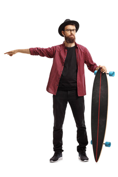 Full length portrait of a male hipster holding a longboard and pointing in one direction isolated on white background - Zdjęcie, obraz