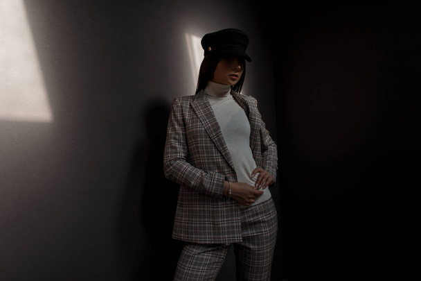 Stylish beautiful young woman in trendy hat and stylish vintage suit posing on a dark background with light - Fotó, kép