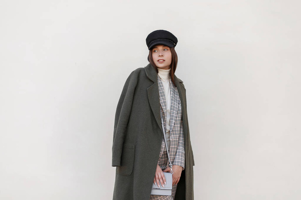 Fashionable woman with a hat in a stylish green coat with a checkered fashionable suit and bag posing near the wall - Foto, imagen