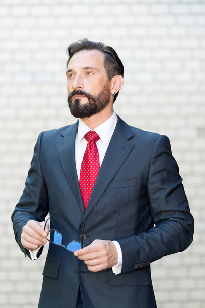 Bearded elegant man in dark suit thoughtfully looking into the distance while standing with his glasses against the white brick wall - 写真・画像