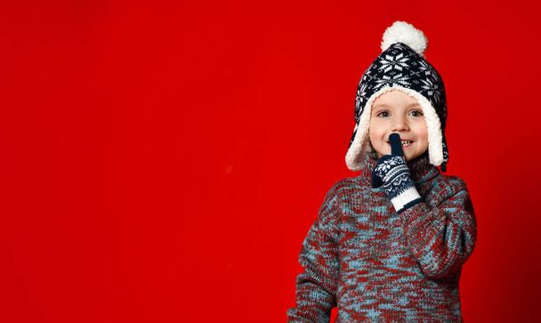 child boy in knitted hat and sweater and mittens making silence gesture  over colorful red  background. - Fotoğraf, Görsel