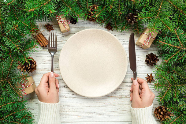 bovenaanzicht meisje houdt vork en mes in de hand en is klaar om te eten. Lege plaat ronde keramische op houten Kerstmis achtergrond. schotel concept van de diner van de vakantie met Nieuwjaar decor. - Foto, afbeelding