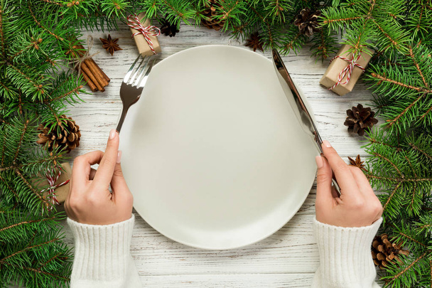 дівчина зверху дивиться, тримає виделку і ніж в руці і готова їсти. Порожня тарілка круглої кераміки на дерев'яному різдвяному фоні. концепція святкової страви з новорічним декором
. - Фото, зображення