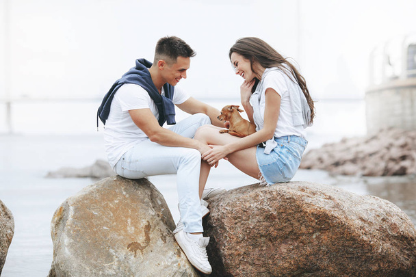 Young beautiful positive couple with small dog sitting on stone by sea. Lifestyle and pets - Photo, Image