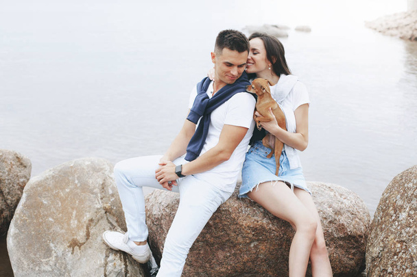 Young beautiful positive couple with small dog sitting on stone by sea. Lifestyle and pets - 写真・画像