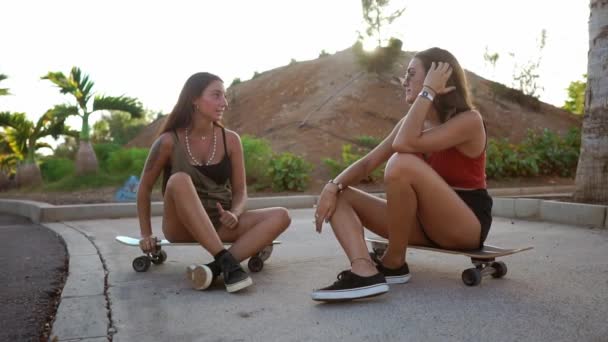 Dos chicas en Skate Park se sientan en las tablas y hablan sonriendo y riéndose de los chistes bajo la luz del atardecer. Longboard conversación amigos
 - Imágenes, Vídeo