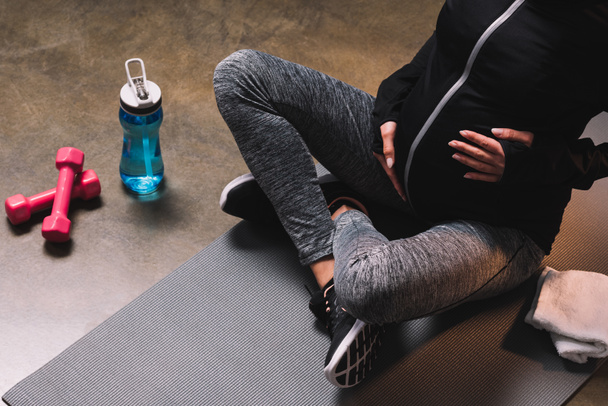 Cropped view of pregnant sporty woman sitting on the sportive mat and sport bottle beside - Фото, изображение