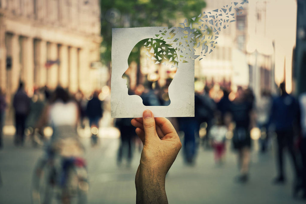 Hand holding a paper sheet with human head icon broken into pieces over a crowded street background. Concept of memory loss and dementia disease. Alzheimer's losing brain and memory function. - Photo, Image